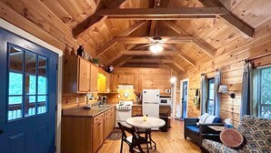 A cozy cabin interior with wooden walls and ceilings, a kitchen with basic appliances, a dining area with a table and chairs, and a living area with a sofa and a log. - Cozy Blue Skies Cabin - walk to Chimney Rock & Lake Lure (Lake Lure)