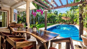 Een zwembadterras met een houten tafel en stoelen, een pergola met plafondventilator en uitzicht op een zwembad omringd door weelderige begroeiing.