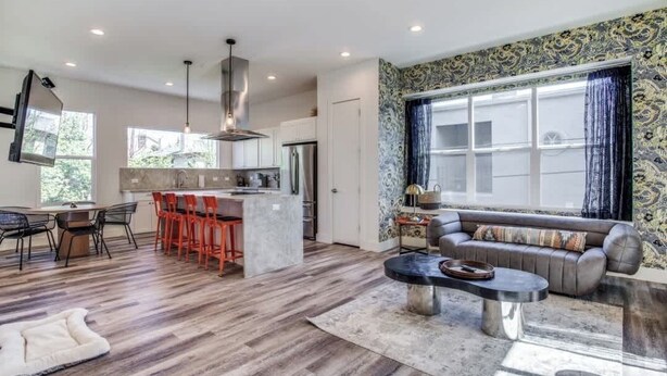 A modern kitchen with a central island, a dining area with red chairs, a living room with a sofa and coffee table, and a large window with patterned curtains.