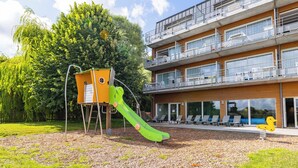 Un edificio moderno con varios balcones, un parque infantil con tobogán y un gran árbol al fondo.