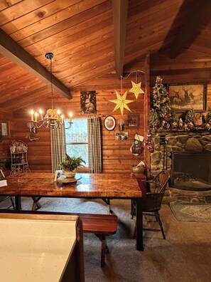 L’intérieur chaleureux d’un chalet avec une table à manger en bois, des bancs, un lustre, une cheminée et des décorations de fêtes.