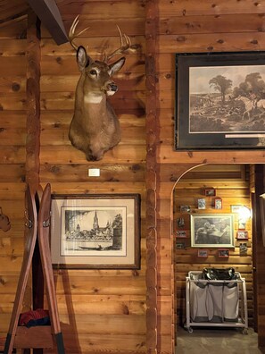 Interior de una cabaña de madera con un venado montado como trofeo, fotografías históricas enmarcadas y una escalera.