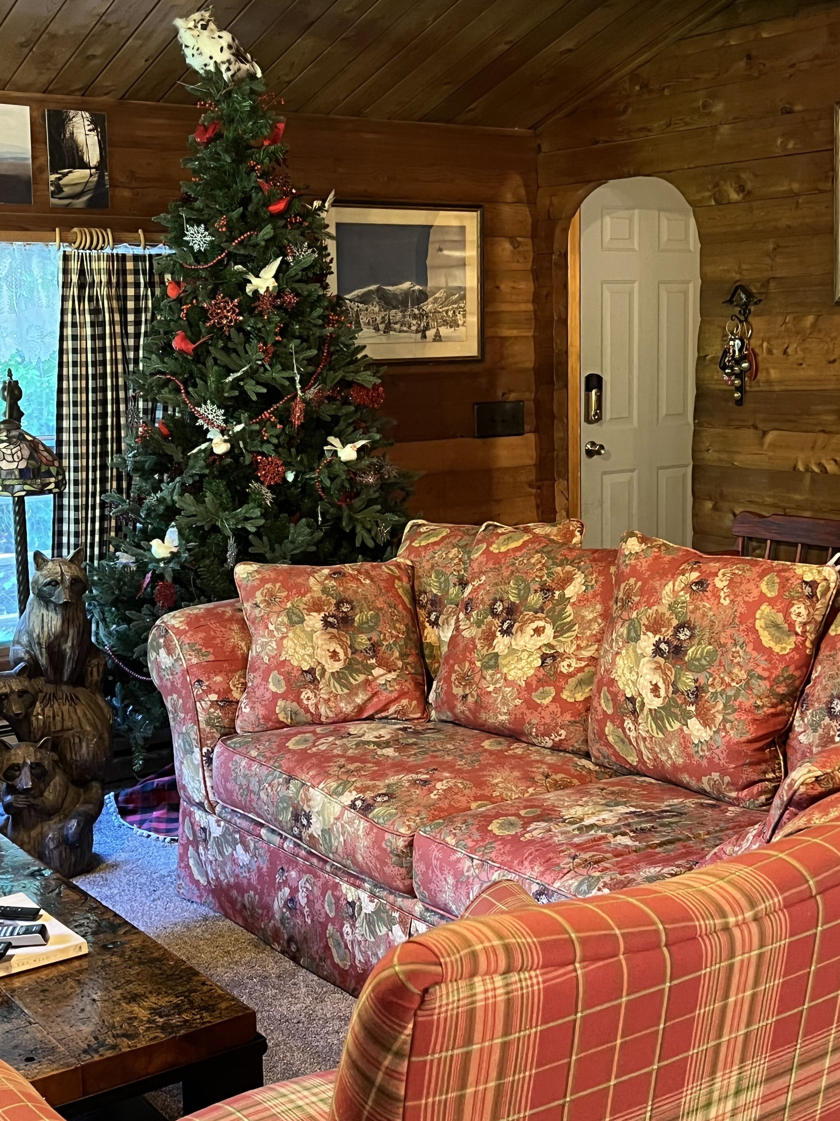 A cozy cabin interior with a wooden ceiling, a Christmas tree, a floral sofa, and a rustic decor.