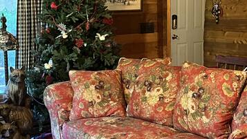 A cozy cabin interior with a wooden ceiling, a Christmas tree, a floral sofa, and a rustic decor.
