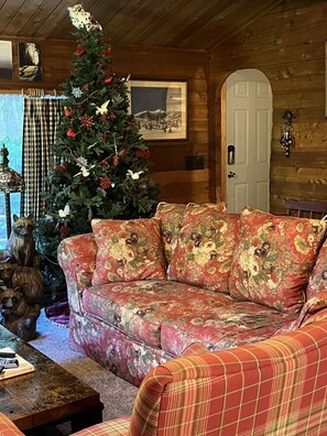A cozy cabin interior with a wooden ceiling, a Christmas tree, a floral sofa, and a rustic decor.