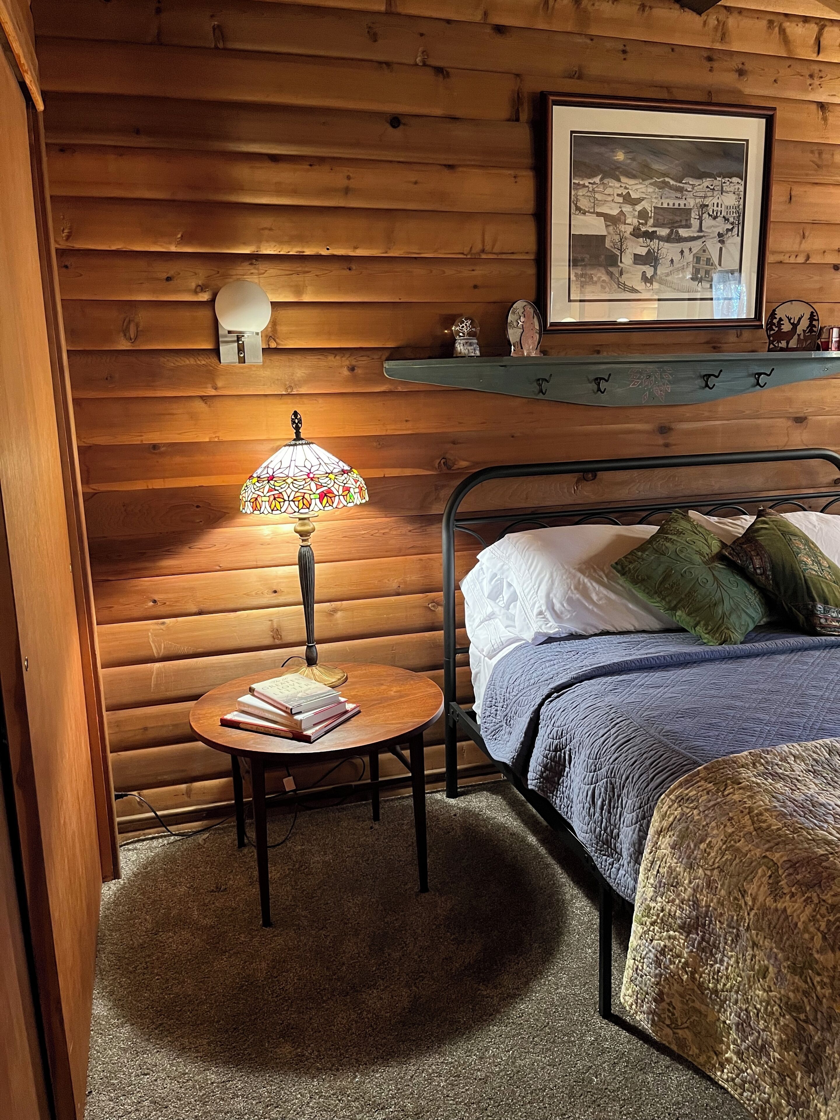 A cozy room with a wooden log wall, a bed with a blue quilt, a round table with a lamp, and a framed picture above the bed.