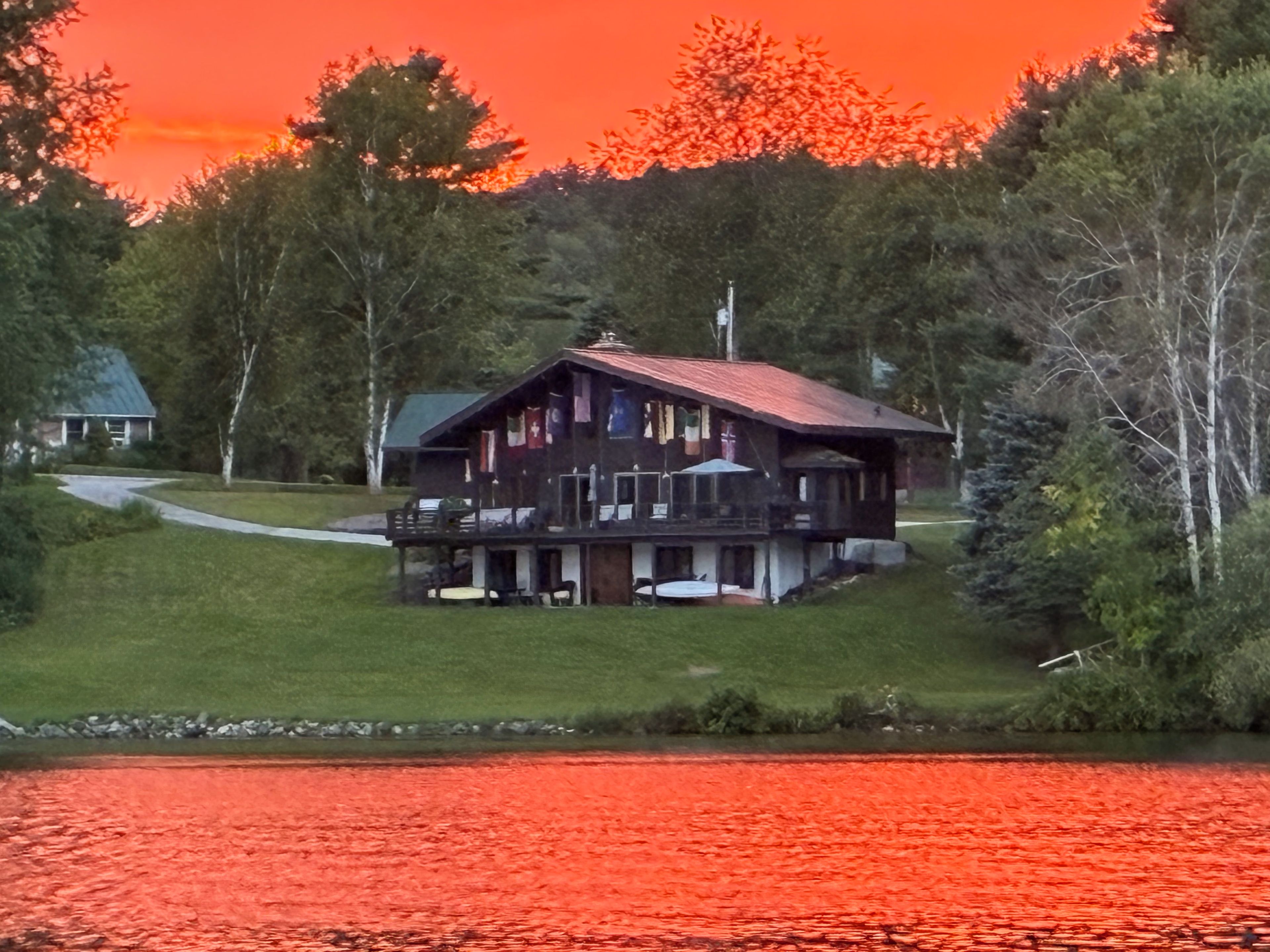 A two-story cabin with a red roof, situated on a grassy hill overlooking a body of water.