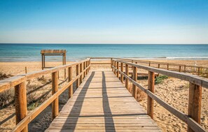 Ein hölzerner Steg führt zu einem Strand mit einer Bank und einem Rettungsschwimmerstand.