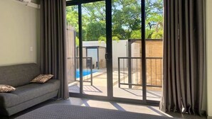 Une chambre d’hôtel moderne dotée d’un canapé, d’un grand lit et d’une porte-fenêtre coulissante donnant sur un espace extérieur avec une piscine et une terrasse en bois.