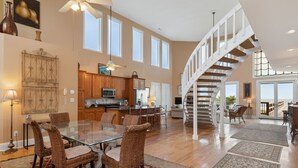 A spacious living area with a glass dining table, wicker chairs, a kitchen with wooden cabinets, and a spiral staircase. - All Occasion House! Grand Oceanfront Home, Direct Beach Access, Large Ocean View Windows (Folly Beach)
