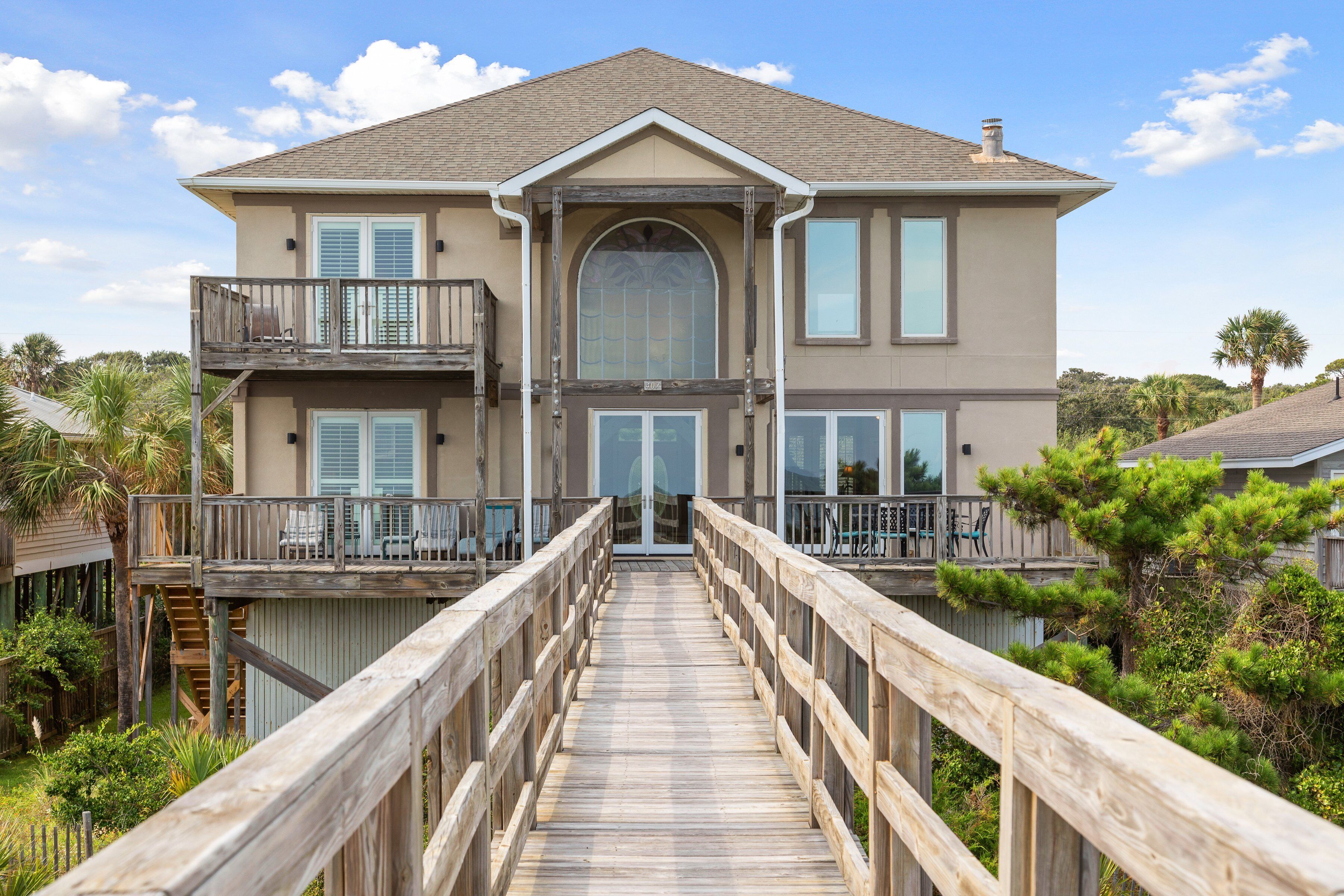 A two-story house with a wooden deck leading to the front door, surrounded by greenery.