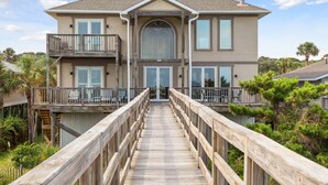 A two-story house with a wooden deck leading to the front door, surrounded by greenery. - All Occasion House! Grand Oceanfront Home, Direct Beach Access, Large Ocean View Windows (Folly Beach)
