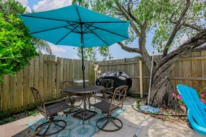 A patio with a table and chairs, a blue umbrella, and a barbecue grill.