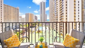 Ein Balkon mit zwei Stühlen, einem kleinen Tisch mit einer Pflanze und zwei Tassen, sowie Blick auf Hochhäuser und einen Poolbereich.