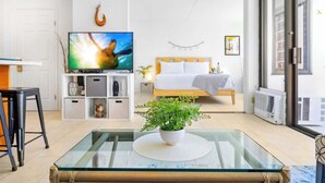 Ein modernes Wohnzimmer mit einem Glastisch, einem Bett mit weißem Kopfteil, einem Fernseher und einem großen Fenster.