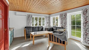 A cozy living room with a wooden ceiling, a black sofa, a wooden coffee table, and a view of a garden through large windows.