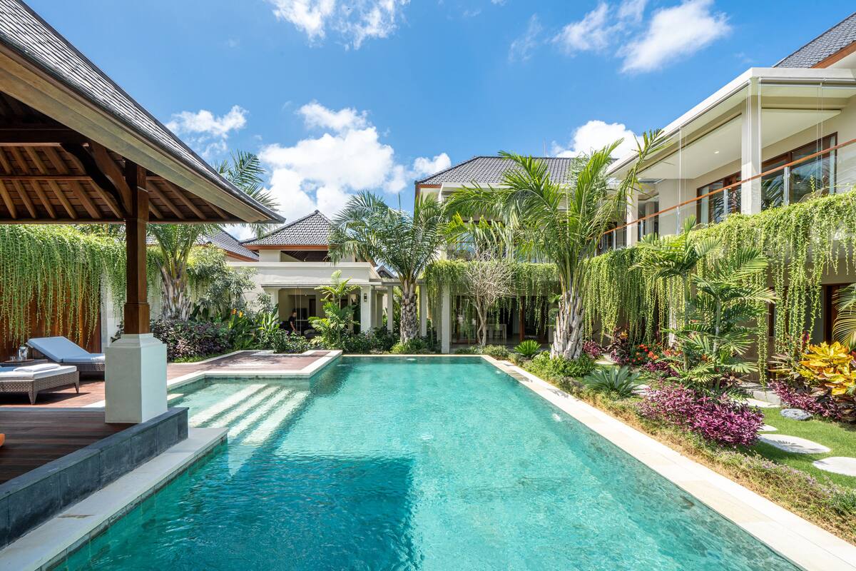 Uma piscina com deck de madeira e espreguiçadeiras, cercada por plantas tropicais e uma casa moderna com telhado de azulejos.