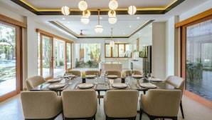 A dining area with a large table set for six, surrounded by chairs, with a view of a kitchen and outdoor greenery. - Villa Reillo (Canggu)