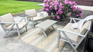 Een terras buiten met een zithoek bestaande uit een bank en twee stoelen, een kleine tafel en een decoratieve plant met roze bloemen.