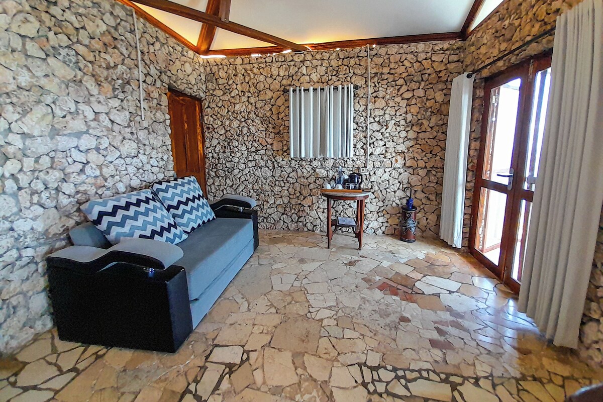 A room with a stone wall, a wooden ceiling, a sofa with patterned cushions, a small table with a kettle, and a window with curtains.