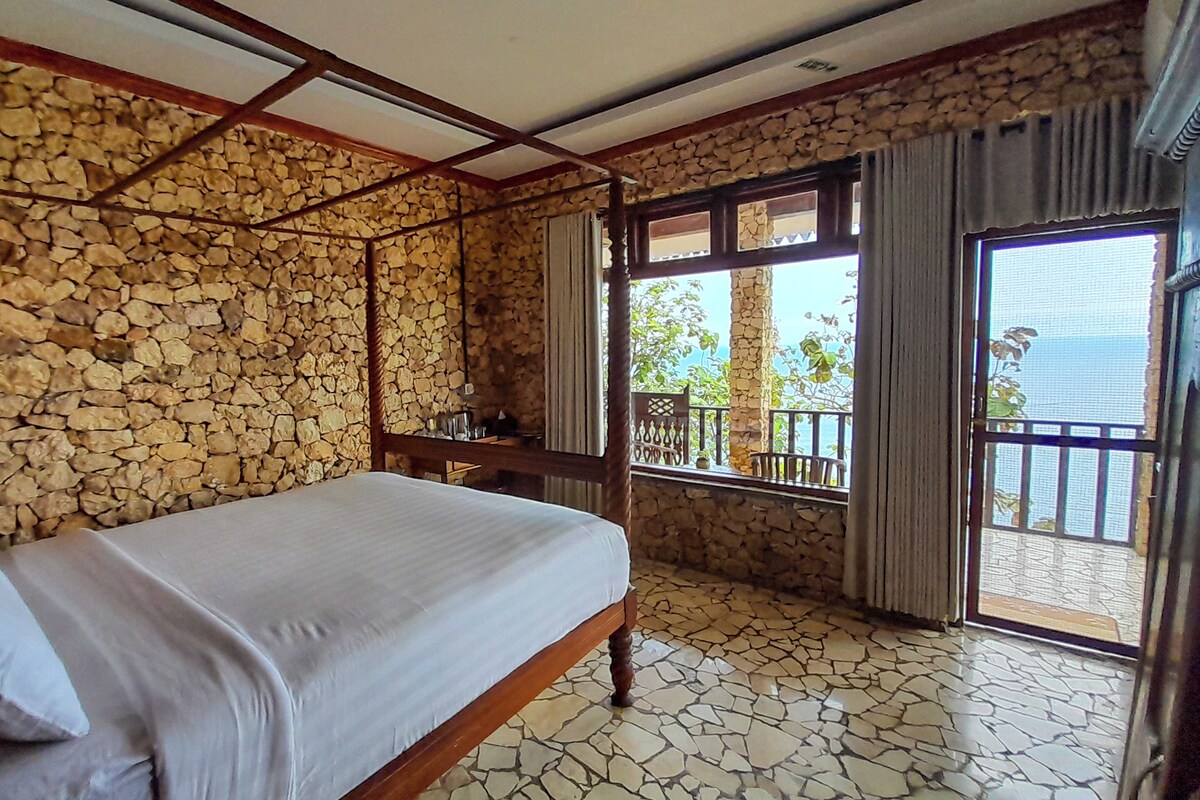 A room with a stone wall, a bed, a wooden desk, and a balcony with a view.