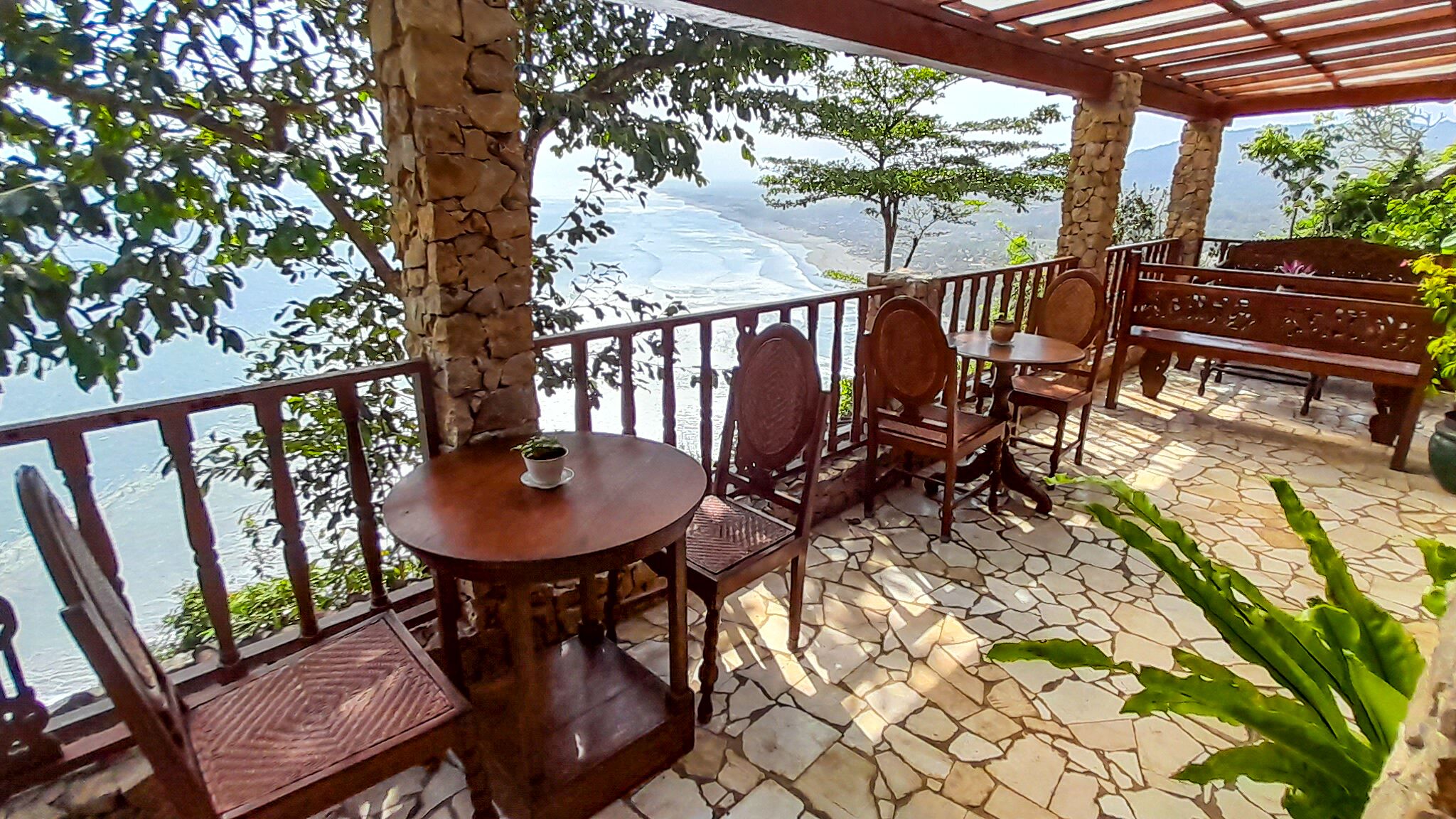 A covered outdoor seating area with stone flooring, wooden furniture, and a view of a body of water.