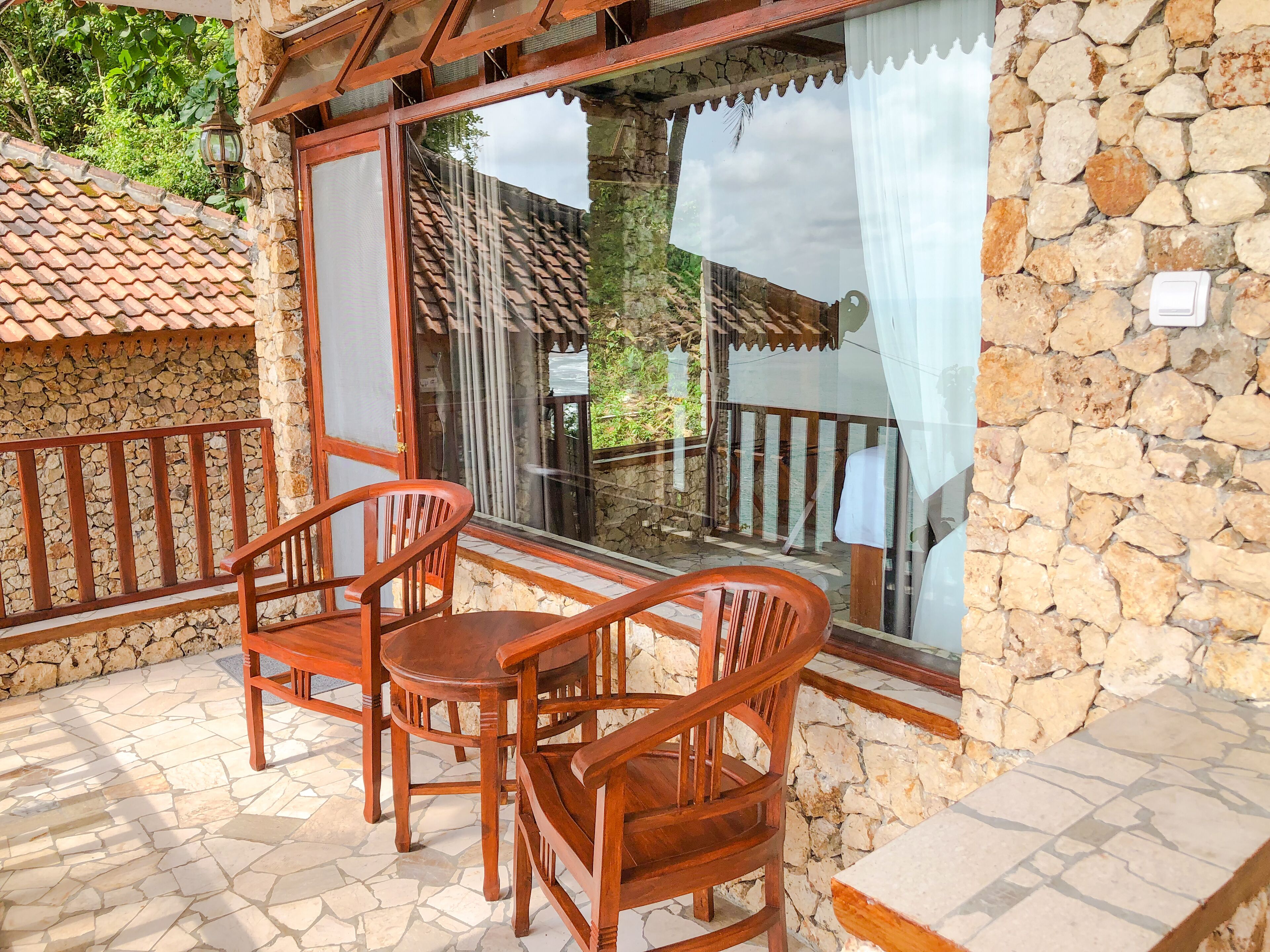A stone-walled balcony with wooden furniture and a view of a tiled roof and greenery.