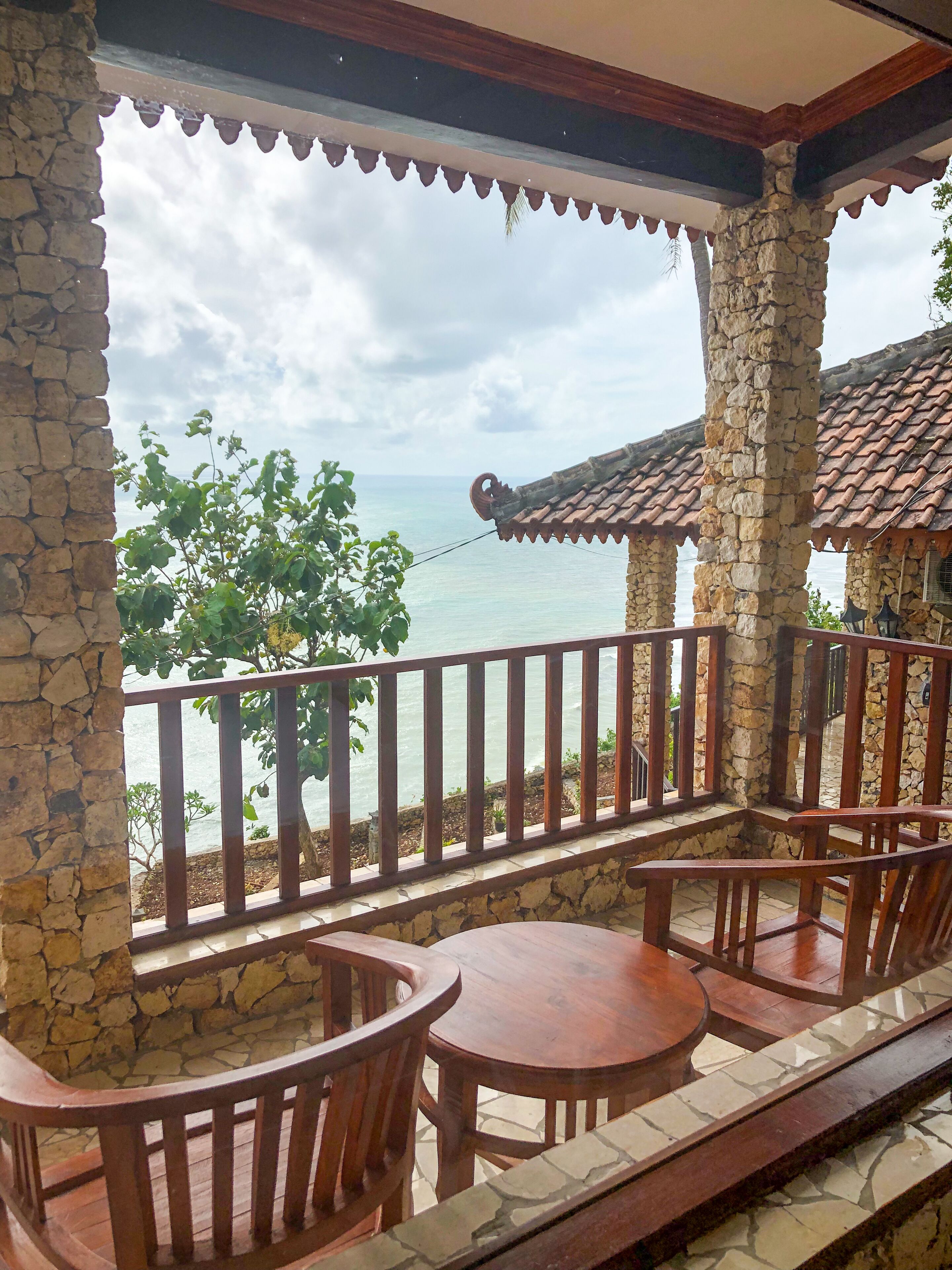 A stone-walled balcony with wooden furniture, overlooking a body of water and a building with a tiled roof.