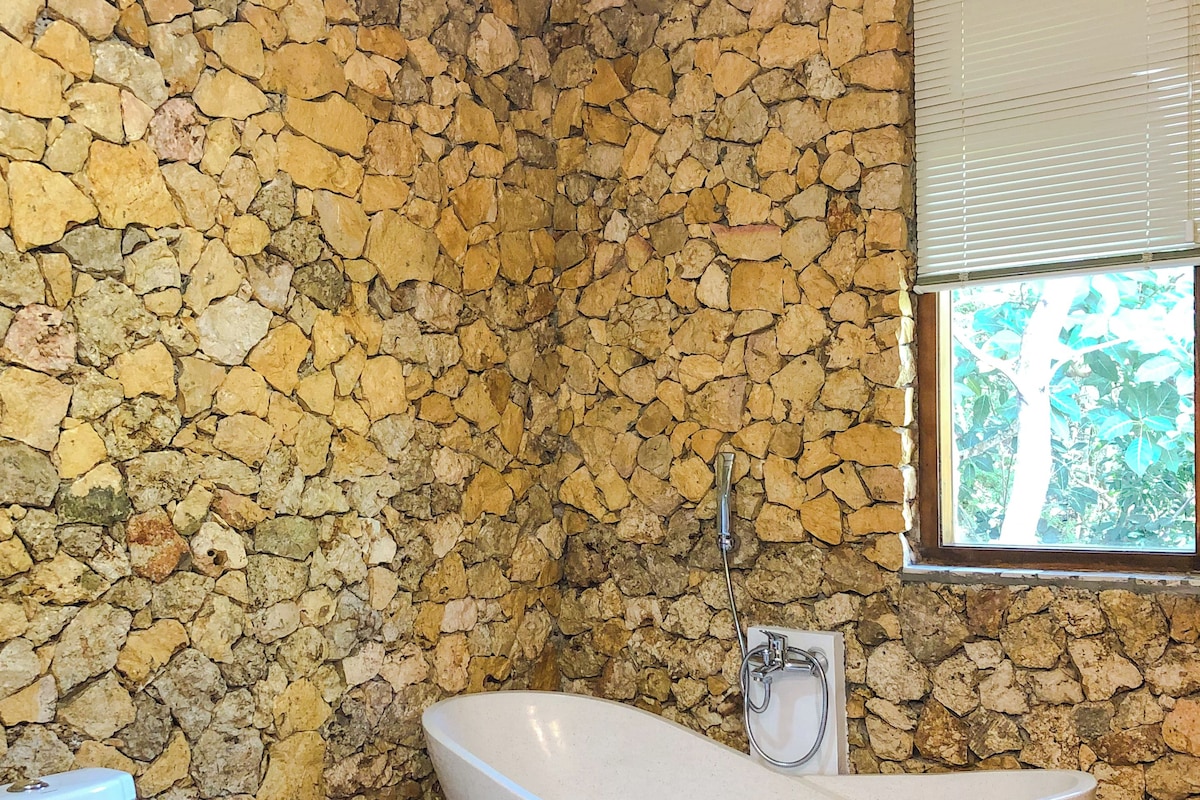 A bathroom with a stone wall, a freestanding bathtub, a toilet, and a window with blinds.