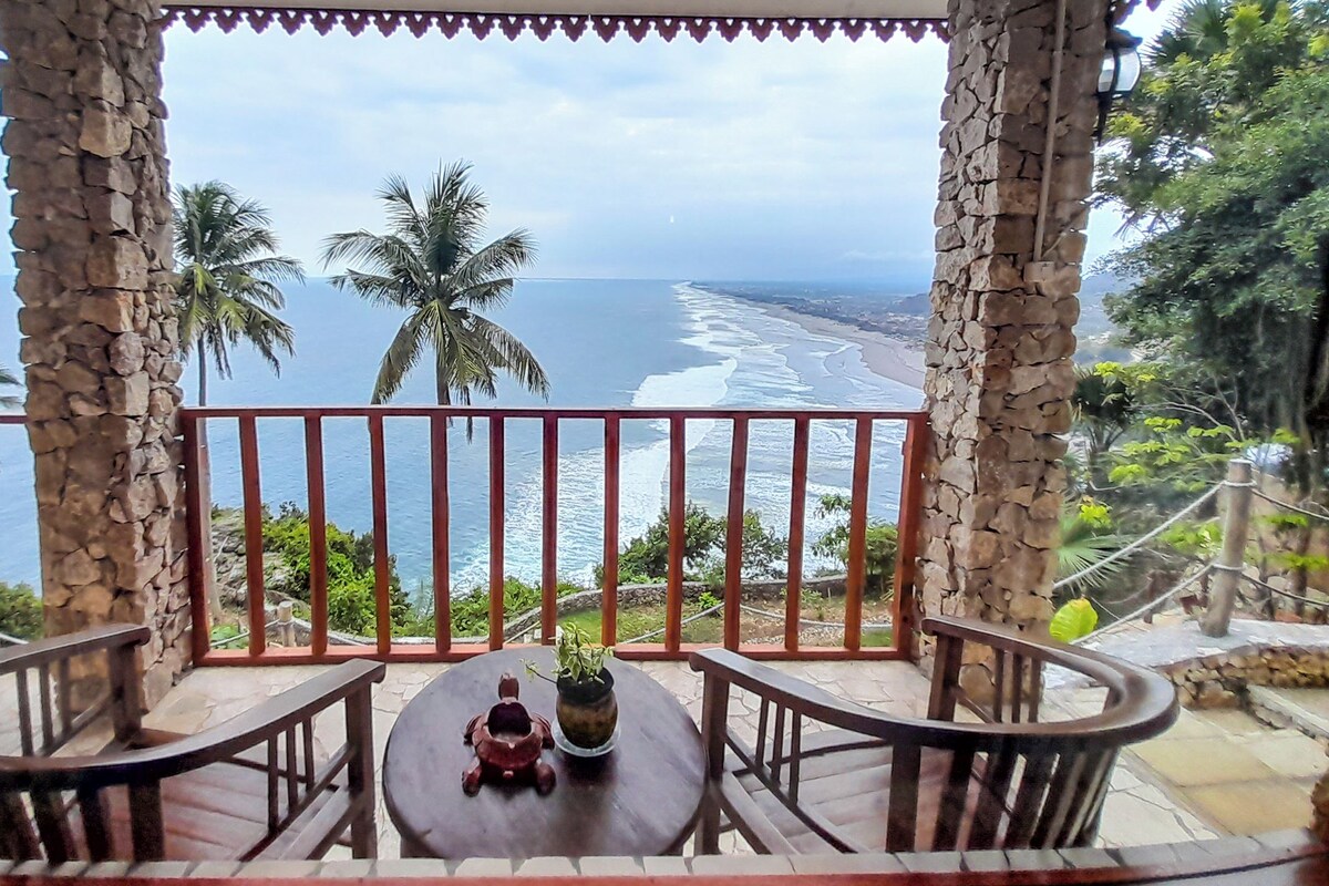 A balcony with a view of the ocean, stone pillars, and a small table with a plant.