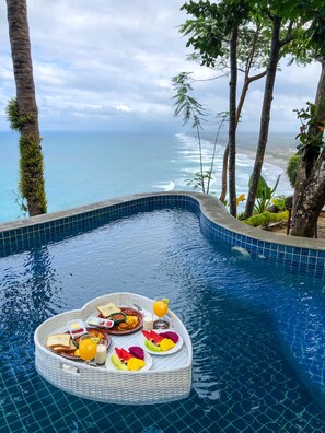 A pool with a breakfast tray containing toast, fruit, and juice, surrounded by trees and overlooking a beach. - Edge Resort Yogyakarta (Giricahyo)