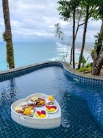 Piscina com bandeja de café da manhã contendo torradas, frutas e suco, cercada por árvores e com vista para uma praia.