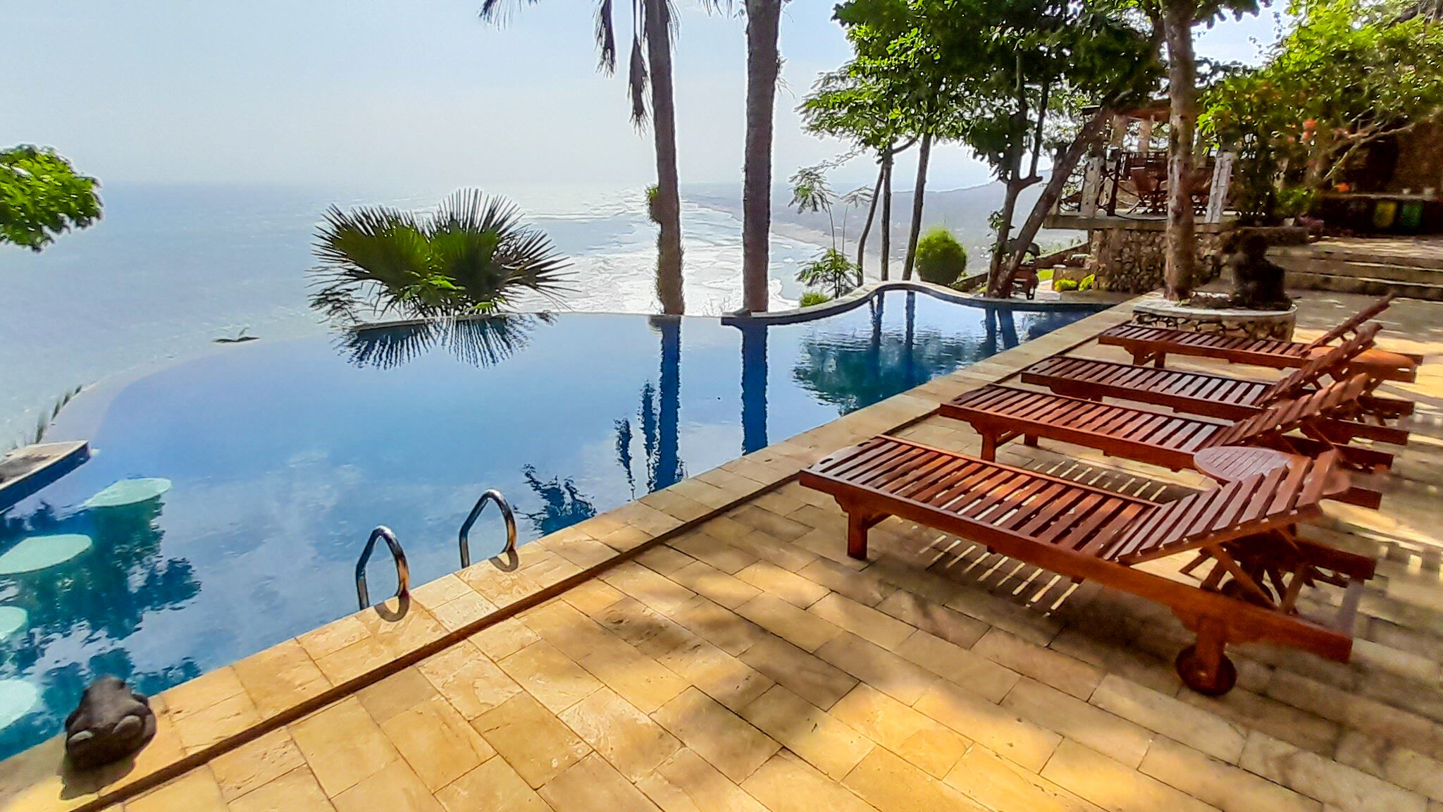 An infinity pool with wooden loungers, a clear view of the ocean, and tropical trees.