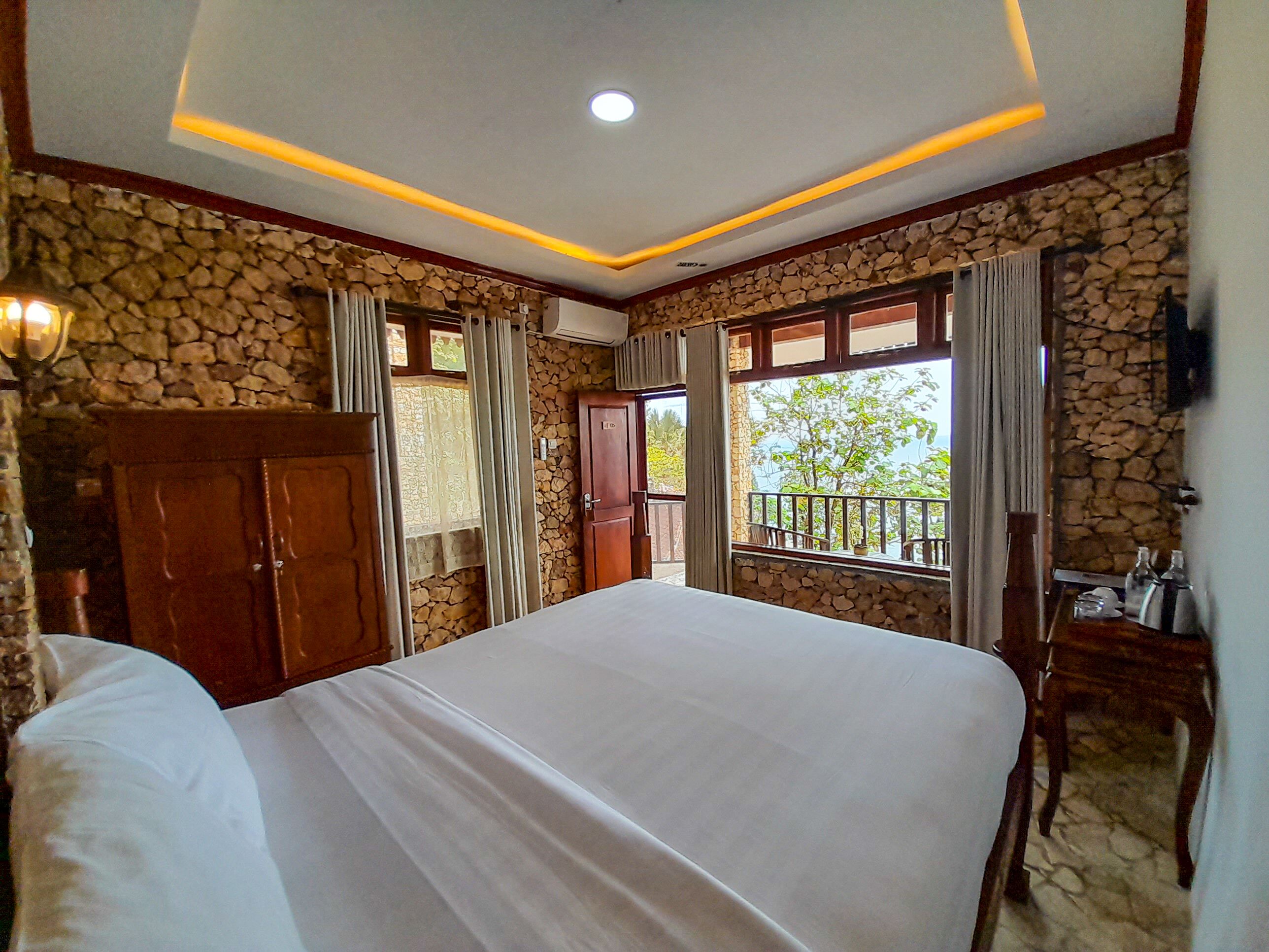 Un dormitorio con una cama grande, paredes de piedra, un balcón con barandilla y un escritorio de madera con una silla.