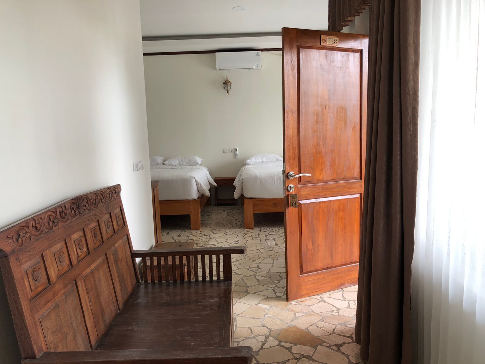 A room with a wooden bench, two beds with white linens, a stone-tiled floor, and a wooden door with a number plate.