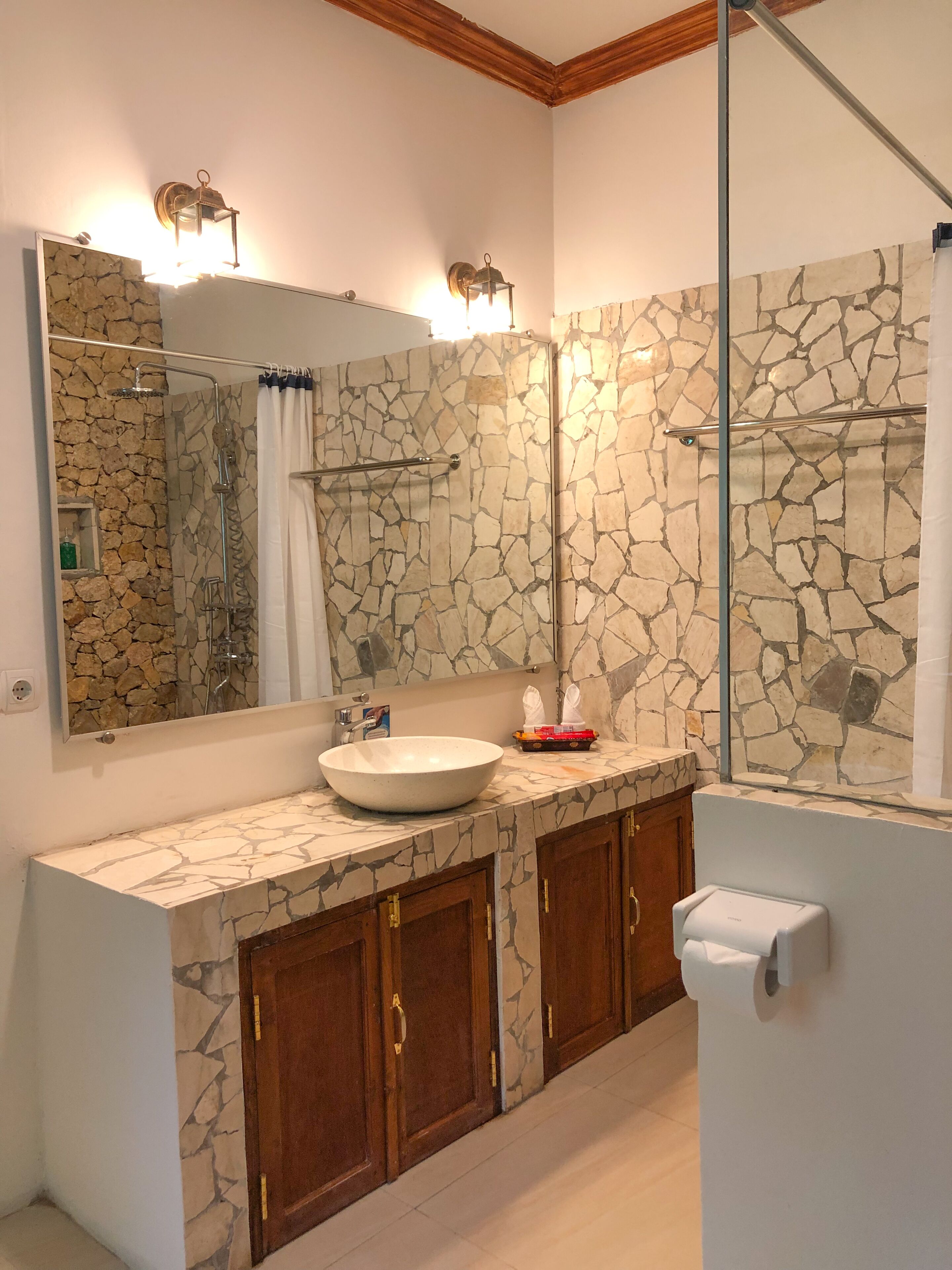 Une salle de bain avec un mur décoré d’une mosaïque en pierre, un grand miroir, un lavabo, des meubles en bois et un espace douche.