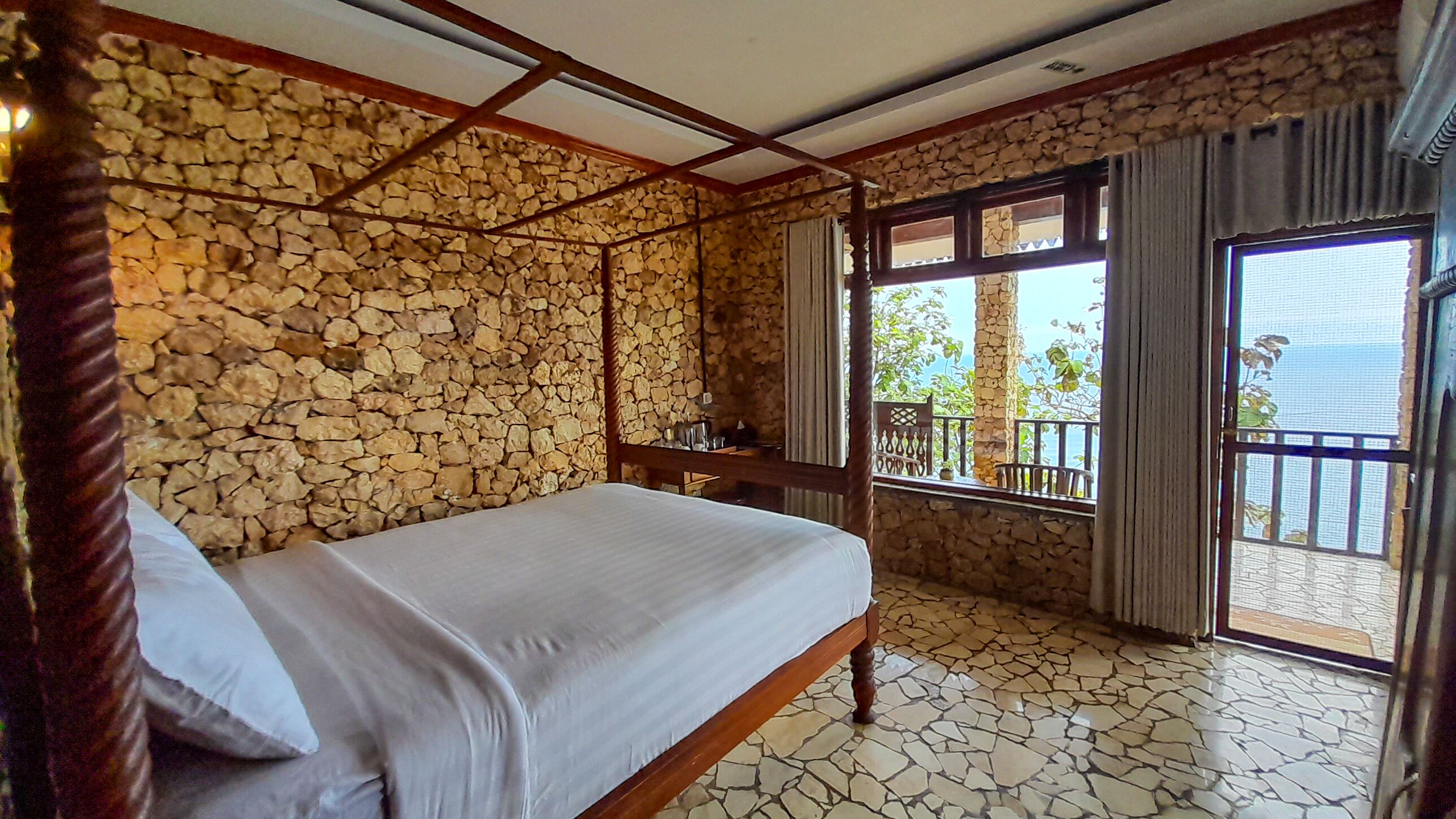 Ein Zimmer mit Steinwand, einem Bett, einem Holztisch und einem Balkon mit Blick.