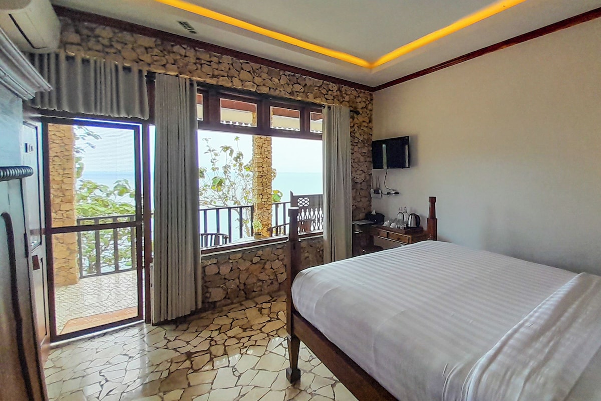 A hotel room with a bed, a desk, a TV, and a balcony with a view of the sea.