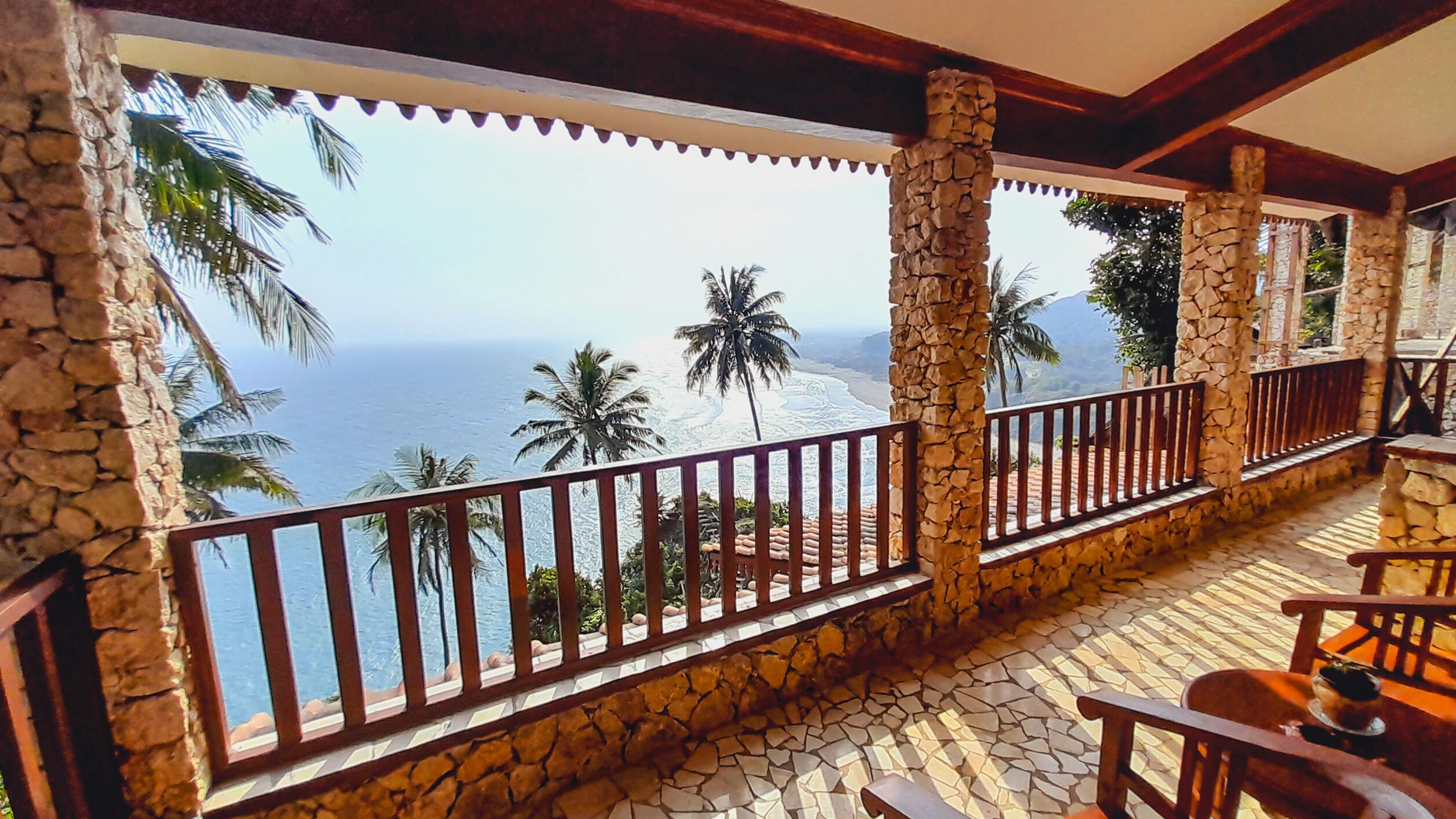 A balcony with stone pillars, a tiled floor, and a view of the ocean and palm trees.