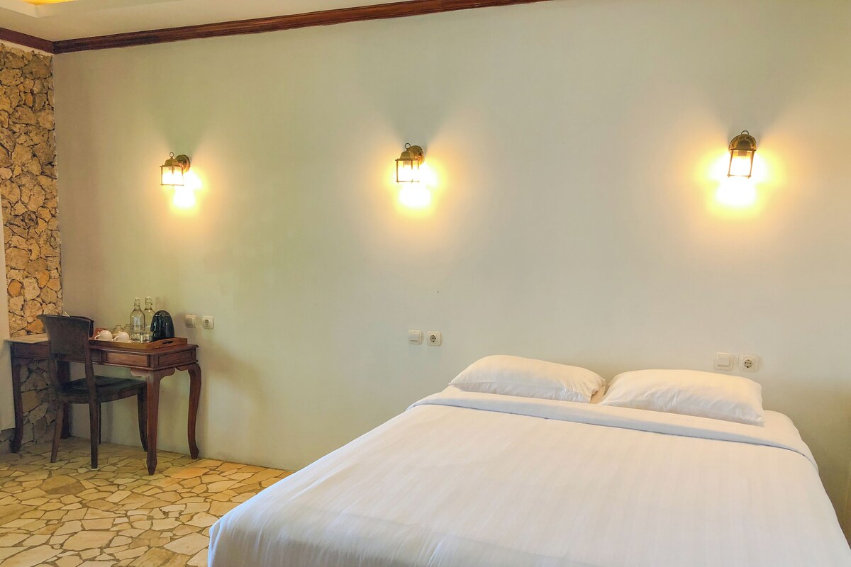 A hotel room with a bed, two wall-mounted lights, a desk with a chair, and a stone wall.