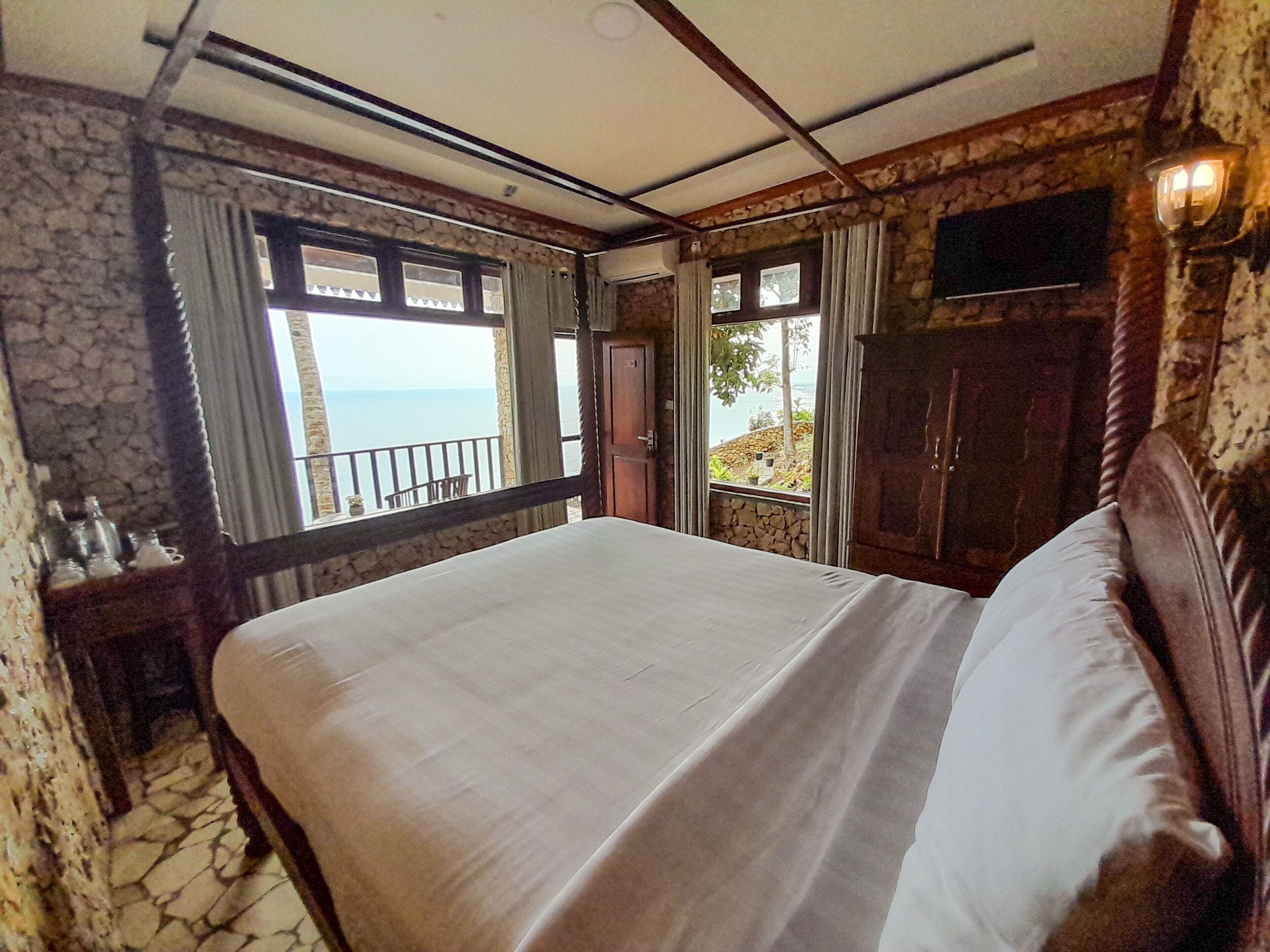 A bedroom with a large bed, stone walls, a balcony with a view of the sea, and a wooden wardrobe.