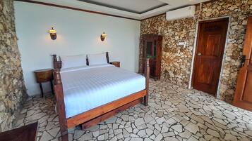 A bedroom with a large bed, two wall-mounted lights, a stone wall, a wooden door, and a stone-tiled floor.
