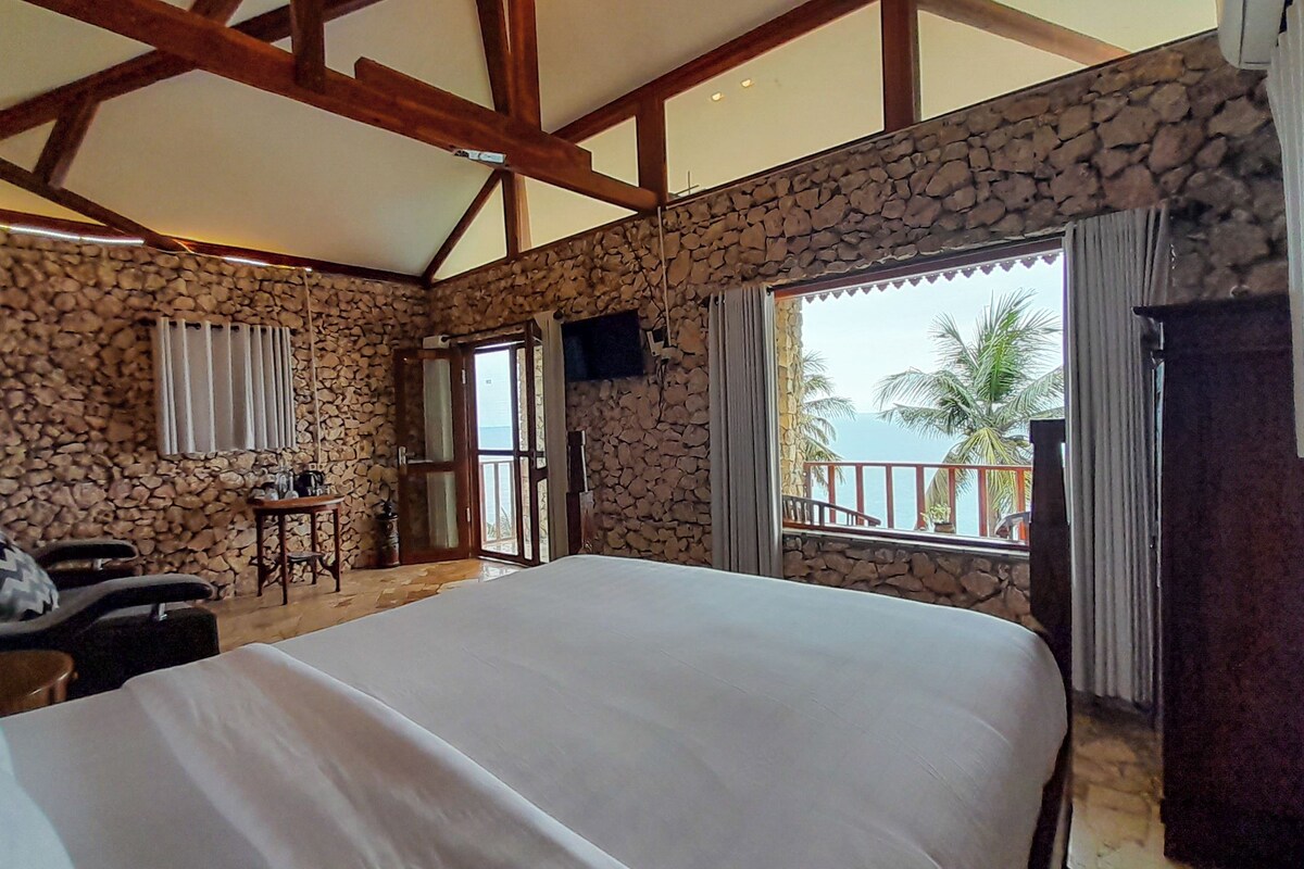 A bedroom with a stone wall, a bed, a small table, and a view of the sea.