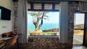 A room with a stone wall, a window with curtains, a wooden desk, and a view of the sea. - Edge Resort Yogyakarta (Giricahyo)