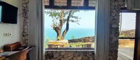 A room with a stone wall, a window with curtains, a wooden desk, and a view of the sea.