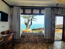 Quarto com parede de pedra, janela com cortinas, escrivaninha de madeira e vista para o mar.