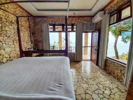 Quarto com parede de pedra, uma cama, uma escrivaninha e vista para o mar através de uma porta de varanda.