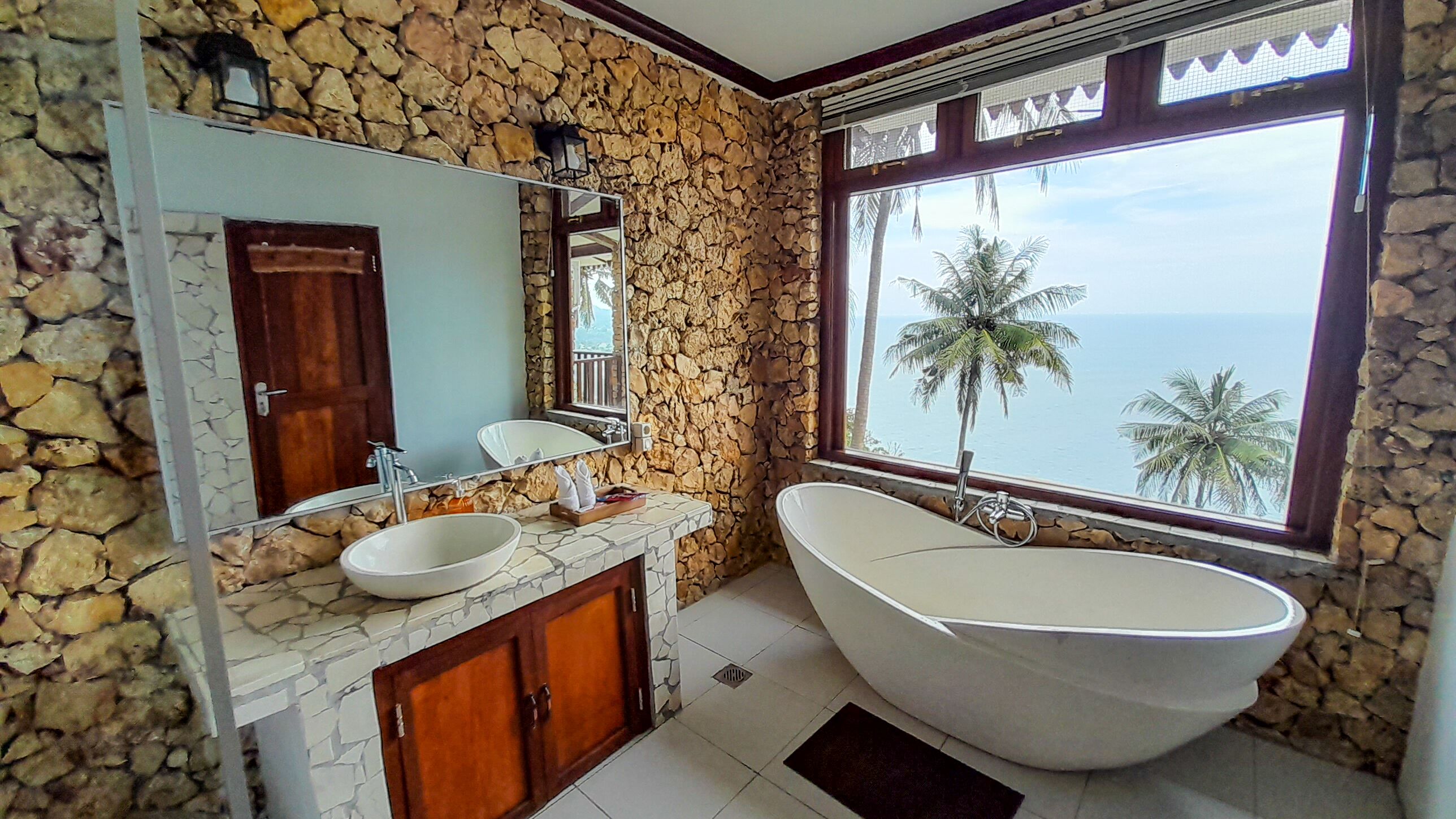 A bathroom with a stone wall, a large window with a view of palm trees and the sea, a freestanding bathtub, and a vanity with a mirror and sink.