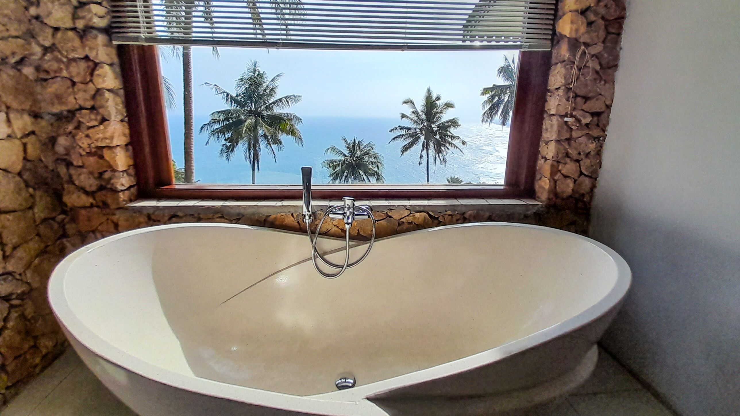 A freestanding bathtub in a bathroom with a stone wall and a window overlooking palm trees and the ocean.