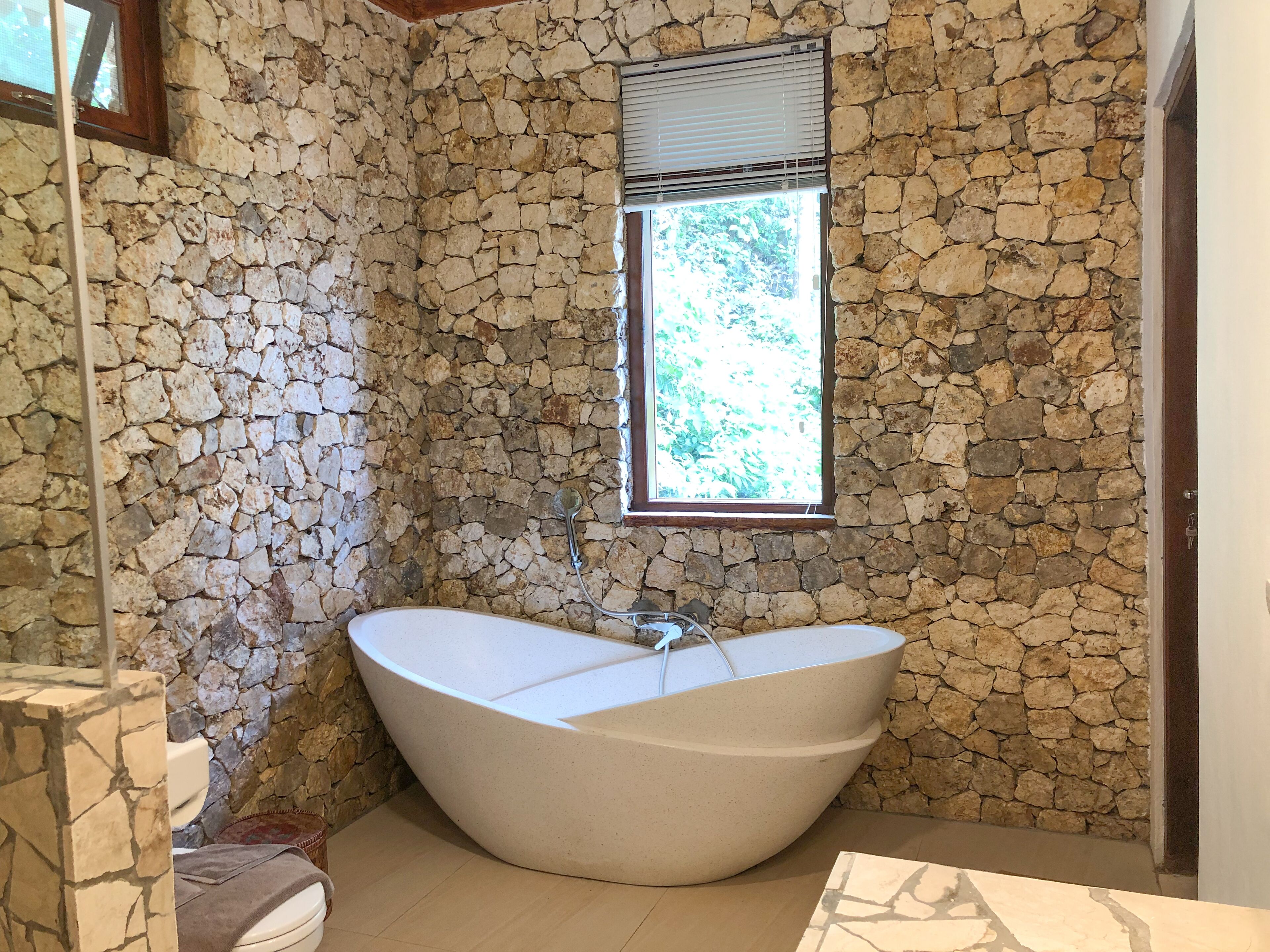 A bathroom with a stone wall, a large bathtub, and a window with blinds.